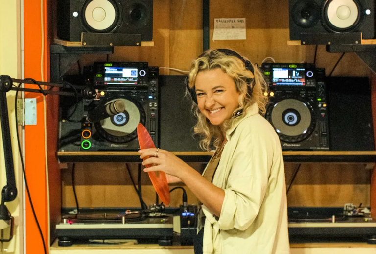 The Porch Sessions founder Sharni Honor smiling with headphones on inside a radio studio booth holding a orange vinyl record next to DJ decks.